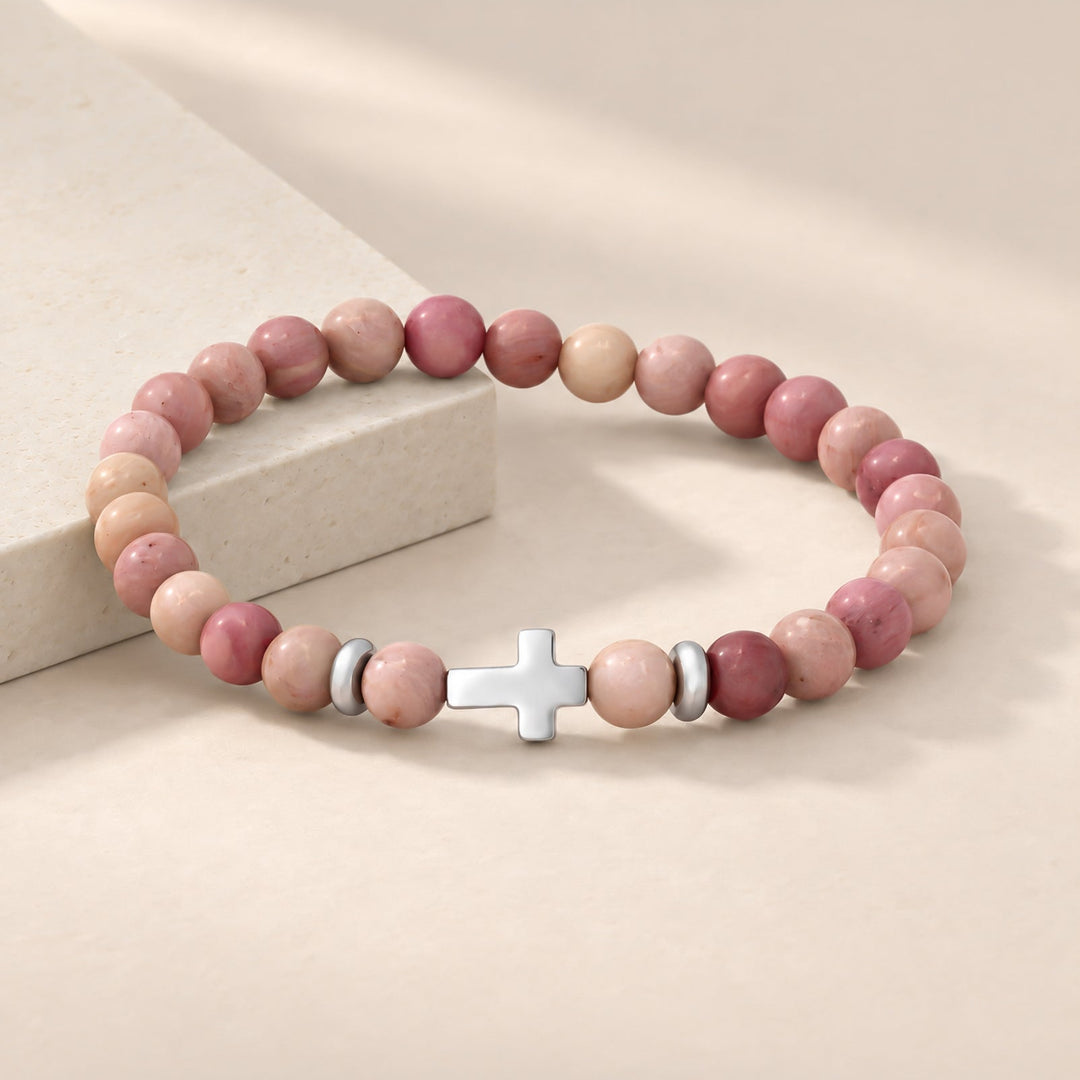A beaded stretch bracelet with pink and peach stone beads, a silver cross charm, and small silver spacer beads, displayed on a warm beige surface next to a light stone block.