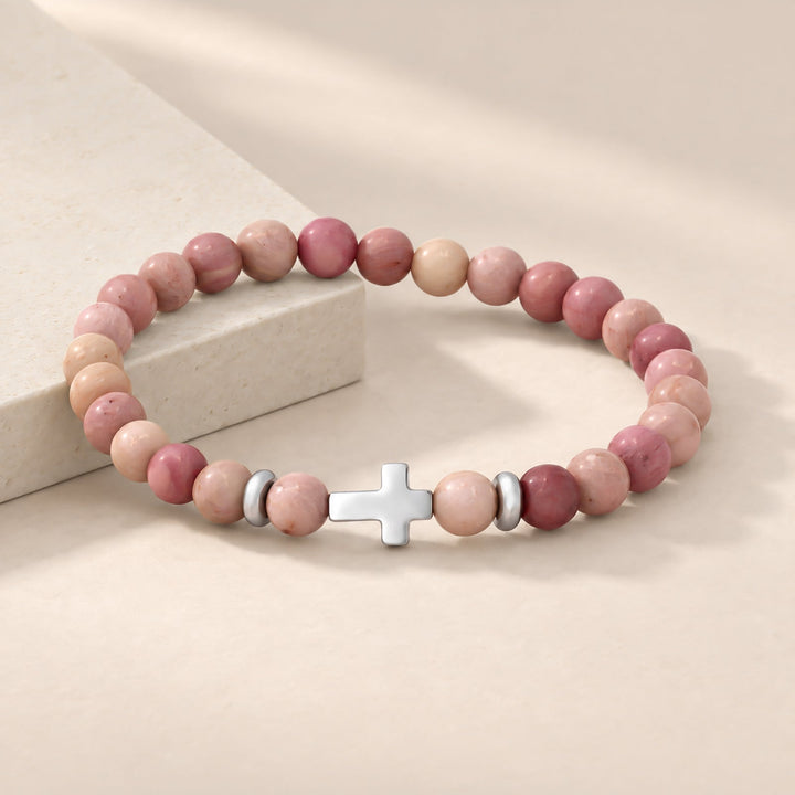 A beaded stretch bracelet with pink and peach stone beads, a silver cross charm, and small silver spacer beads, displayed on a warm beige surface next to a light stone block.