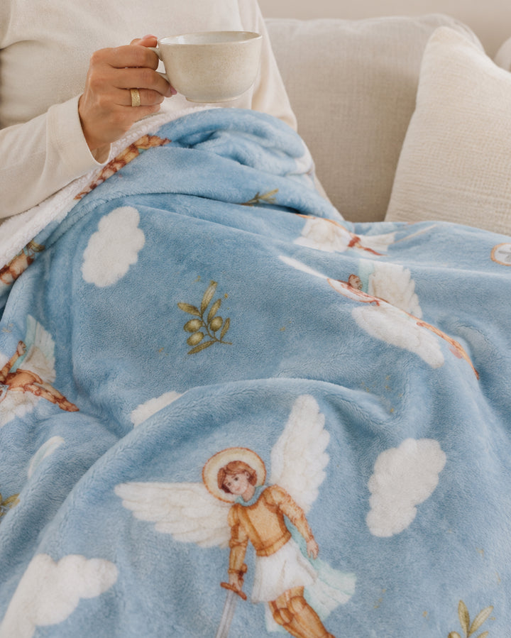 A person sitting on a couch under a plush light blue blanket printed with white clouds and angel illustrations, holding a white mug.